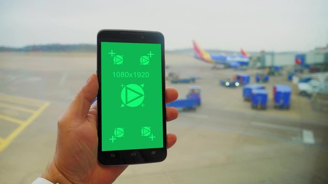 A man holds a green screen smartphone while looking out a window at an airport. Baggage carriers and planes in the distance. Green screen with tracking points for screen replacement.  	