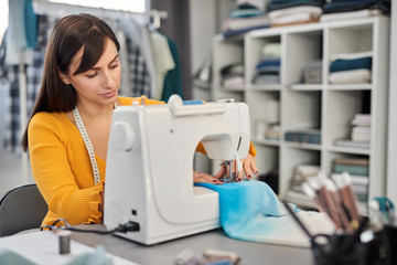 Focused charming caucasian fashion designer sitting in her studio and sewing beautiful evening dress.