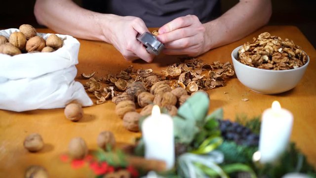 Close up view of male hands cracking walnuts with a nutcracker and putting clean kernels in a bowl. Whole walnuts in a bag, scattered nuts and shells on a table, burning candles on a Christmas wreath