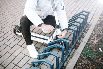 man hangs a cable lock on an electric scooter in a bicycle parking lot. Protection against theft of vehicles.