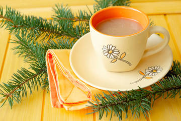 Hot chocolate in orange mug with green branche of pine close up on yellow planks background