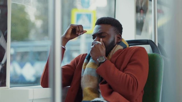 Unhealthy young business guy of african ethnicity suffering cold sneezing allergy messing around on seat driving in tram to work, Bad condition concept.