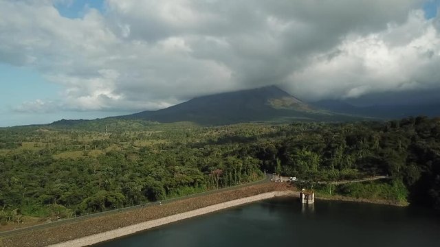 Aerial images of the beautiful landscape on Lake Arenal, Costa Rica, with the Volcano and the lake in the background in a cloudy day, with cars passing by on the highway.
