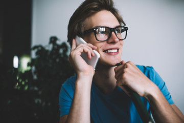 Happy cheerful hipster guy in optical glasses for provide eyes protection rejoicing during cellphone conversation connected to 4g wireless for making phone call, millennial man communicating via app