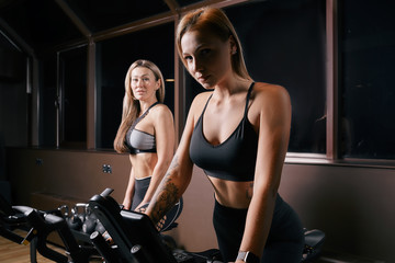 Naklejka na ściany i meble Two attractive sporty women riding exercise bikes during cycling training in gym
