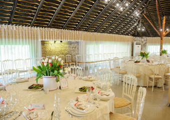 close up photo of a banquet room at a christening party: arranged white round tables with vases with tulips and plates with dishes on it