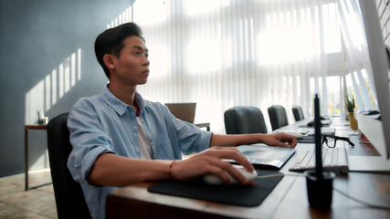 Designer at work. Serious young asian man in casual wear working with computer while sitting at his workplace in the modern office. Creative agency
