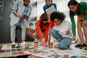 Working on a new project. Group of young interior designers in casual clothes discussing sketches, choosing colors from palettes lying on the floor in the modern office. Creative agency