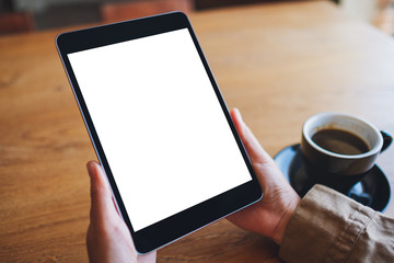 Obraz premium Mockup image of hands holding black tablet pc with blank white screen with coffee cup on wooden table