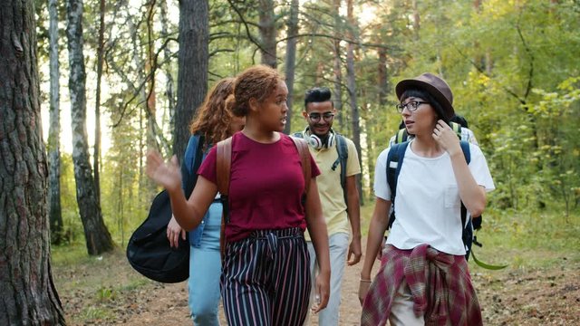 Young cheerful men and women walking in wood doing high-five travelling together talking, people are carrying backpacks and wearing casual clothing.