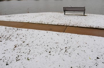Wide shot of snow covering the ground, with a wicker bench  facing a cold pond