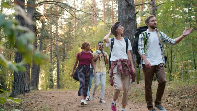 Group of travellers girls and guys with backpacks are walking in forest talking enjoying nature on summer day. Communication, recreation and outdoor activity concept.