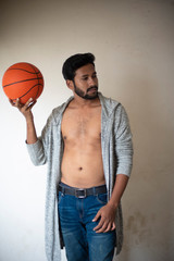 An young tall, dark and handsome Indian Bengali man in a front open western jacket and jeans with basket ball in white background. Indian lifestyle and fashion.