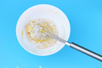 flour with an egg in white bowl and an iron whisk top view of blue background.