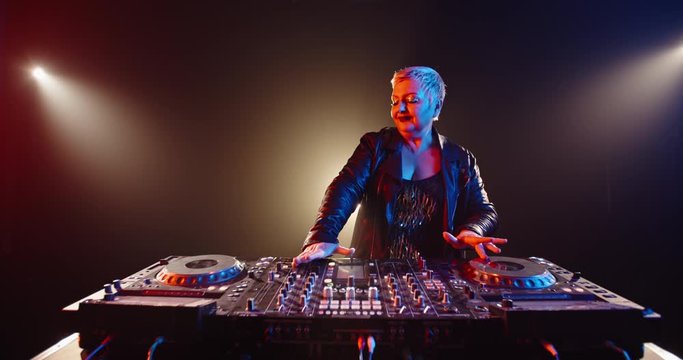 A cool caucasian dj grandma is composing a mix at turntables controller. Authentic mature woman working as disc jockey in a nightclub 4k footage