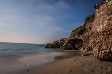 Section of beach in Nerja. Sandy beach on the Spanish coast of Costa del Sol with rocks and stone arch on sunny day with blue sky and clouds. Calm Mediterranean sea with buildings