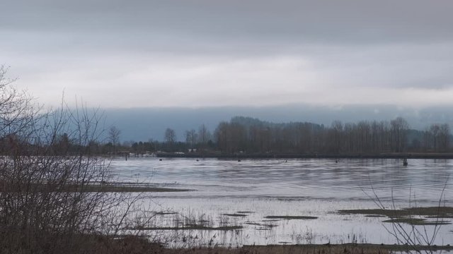 Blue river flowing on a cloudy day with bare branches and dry grass in the foreground and mountains in the background (slow motion)