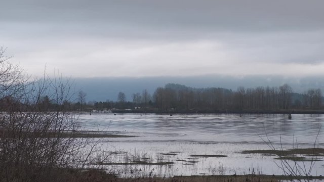 Blue river flowing on a cloudy day with bare branches and dry grass in the foreground and mountains in the background (normal speed)