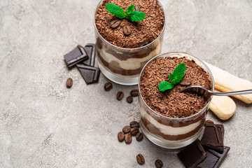 Classic tiramisu dessert in a glass cup, savoiardi cookies and pieces of chocolate on concrete background