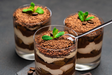 Classic tiramisu dessert in a glass cup and pieces of chocolate on stone cutting board on dark concrete background