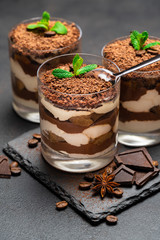 Classic tiramisu dessert in a glass cup and pieces of chocolate on stone cutting board on dark concrete background