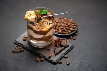 Classic tiramisu dessert in a glass cup and pieces of chocolate on stone cutting board on dark concrete background