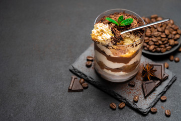 Classic tiramisu dessert in a glass cup and pieces of chocolate on stone cutting board on dark concrete background