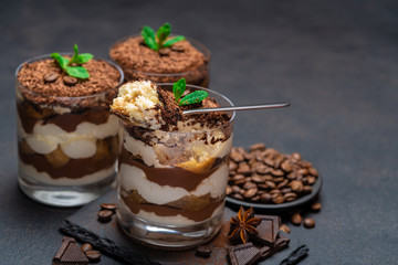 Classic tiramisu dessert in a glass cup and pieces of chocolate on stone cutting board on dark concrete background