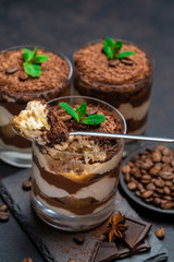 Classic tiramisu dessert in a glass cup and pieces of chocolate on stone cutting board on dark concrete background