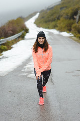 Female runner stretching legs for warming up before running in winter mountain road under the snow. Sporty young woman traning under cold season weather. North of Palencia, Spain.