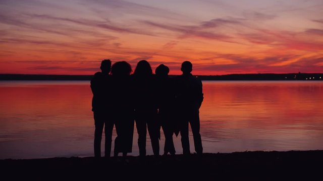 Portrait of happy young people friends watching sun-down on the lake beach standing together hugging talking, beautiful sky and water in the background.