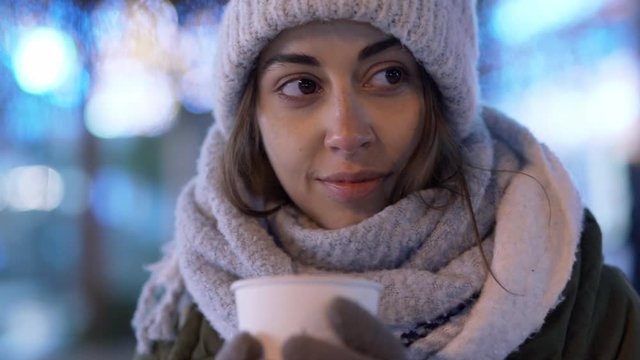attractive young woman in white woolen cap and scarf standing outdoors on illuminated winter city street at evening and drinking coffee from paper cup. woman takes a sip and enjoys the aroma and taste