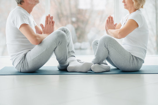 Sports and healthy lifestyle. Croppes image of sporty middle aged couple sitting on the mat in the white fitness gym and doing stretching while looking each other.