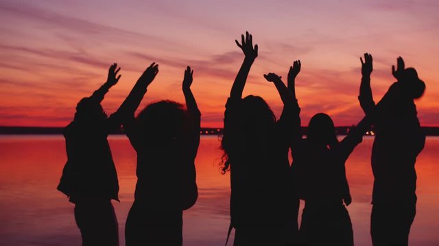 Silhouettes of men and women are dancing on lake shore at night having fun during beautiful simmertime sundown. Lifestyle and outdoor party concept.