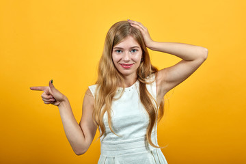 Attractive caucasian blonde young woman keeping hand on head, holding other hand aside, looking pretty over isolated orange background wearing white shirt. Lifestyle concept