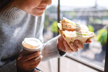 Fotobehang Closeup image of a woman holding and eating whole wheat sandwich and coffee in the morning © Farknot Architect