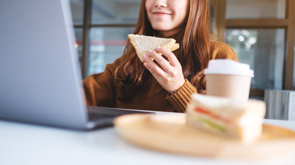 An asian woman holding and eating whole wheat sandwich while working on laptop computer