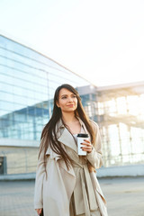  Business woman holding coffee and using phone near office. Portrait of beautiful smiling female with phone, standing outdoors. Phone communication.