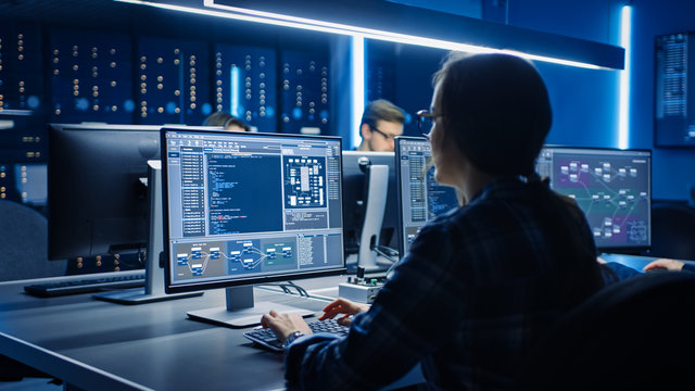 Smart Female IT Programer Working On Desktop Computer In Data Center System Control Room. Team Of Young Professionals Doing Code Programming