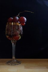 A glass of white sparkling wine with a bunch of grapes on a black background. The glass vintage with gold piping and a white floral pattern.