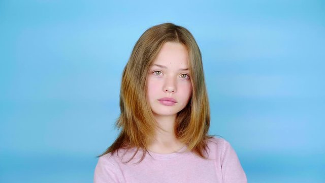 Teen girl in a pink pullover is looking at the camera. Blue background with copy space. Teenager emotions. 4k footage