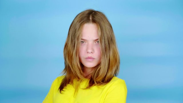 Teen girl in a yellow t-shirt is angry and looking at the camera. Blue background with copy space. Teenager emotions. 4k footage