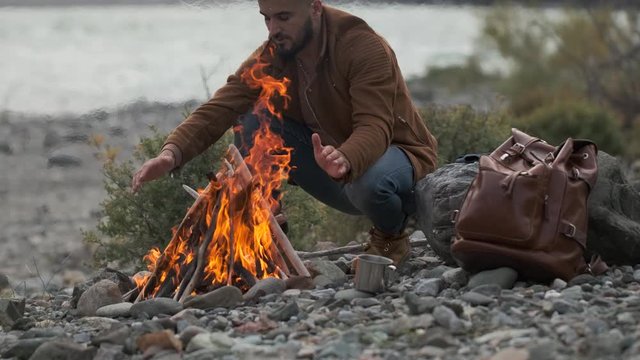 hiker young tourist traveler man enjoying nature, sitting near bonfire at mountain camping camp tent, Active lifestyle traveling fire travel tourism backpacking hiking adventure river. 4 K slow-mo