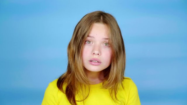 Teen girl in a yellow t-shirt is surprised, opens her mouth and turns her head on camera. Blue background with copy space. Teenager emotions. 4k footage
