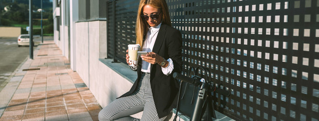 Businesswoman using mobile and drinking coffee sitting on a wall with her scooter next