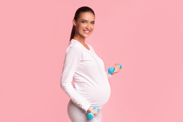 Happy Pregnant Girl Holding Dumbbells Standing On Pink Studio Background