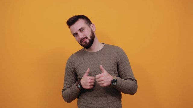 Cheerful unshaven handsome man over isolated orange background looking at camera, keeping thumbs up wearing casual green clothes