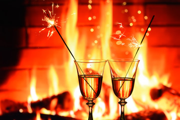 Two glasses with sparkling champagne wine on a burning fire in a fireplace. The cozy atmosphere of winter evenings by the fireplace, the celebration of winter holidays.