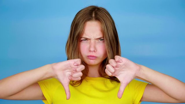 Angry teen girl in a yellow t-shirt is showing thumbs down, dislike and looking at camera. Blue background with copy space. Teenager emotions. 4k footage