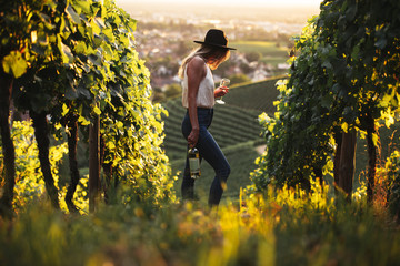 Young caucasian woman standing in the vineyard and posing with a glass of white wine and bottle in hands at sunset. 